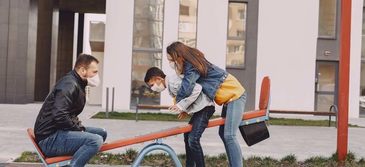 Side view of positive family in trendy casual and medical mask clothes resting near modern building while swinging together on seesaw on urban playground during daytime