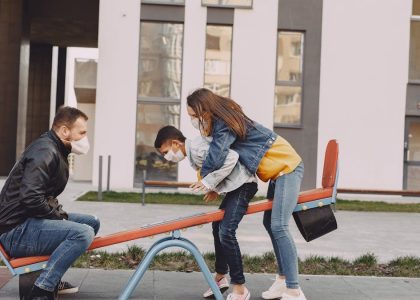 Side view of positive family in trendy casual and medical mask clothes resting near modern building while swinging together on seesaw on urban playground during daytime
